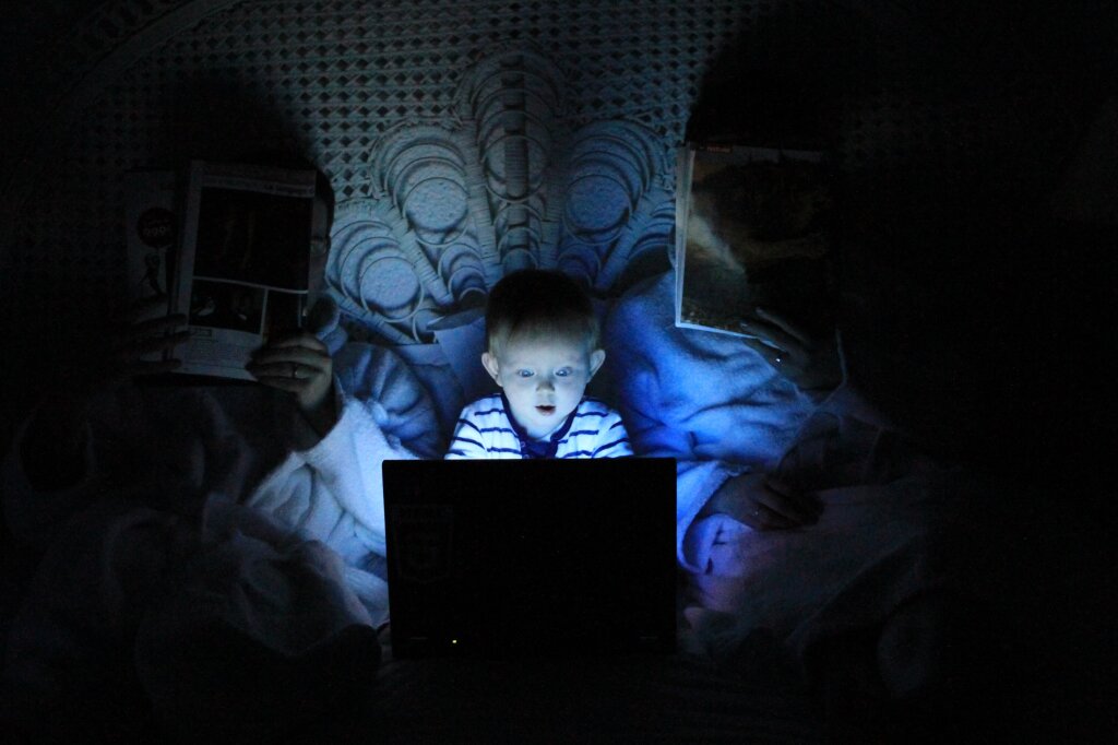 Een jongetje in bed tussen zijn ouders. Een donkere kamer. Laptop op schoot. Deze geeft licht.